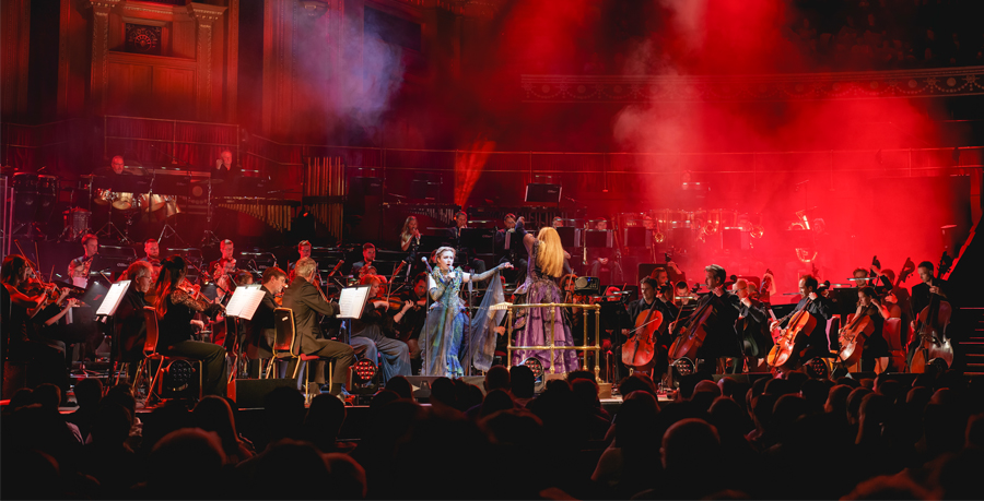A singer and conductor in front of the Orchestra at the Royal Albert Hall, who are under red lighting and smoke effects